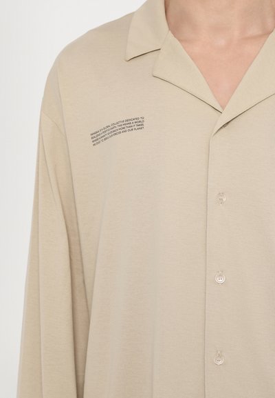Bež lühikeste varrukatega shirt, mis on valmistatud pehmest kangast, klassikalise kraega, nööbid ees ja trükitud tekstiga vasakul küljel.
