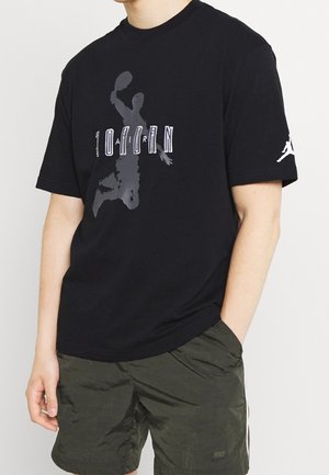 Person iført sort Jordan T-shirt med en grå basketball-silhuet og sorte Nike shorts med hvide striber, hænderne afslappet ned langs siderne.