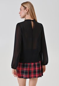Schwarze durchsichtige Bluse mit langen Ärmeln, mit einem Schlüsselloch-Rücken-Detail, kombiniert mit einem roten und schwarzen karierten plissierten Minirock.