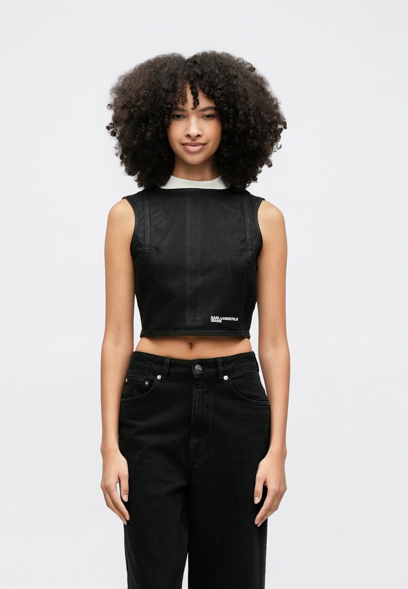 Žena s kučeravými vlasmi v čiernom krátkom tielku bez rukávov a čiernych džínsach s vysokým pásom na jednoduchom svetlom pozadí.