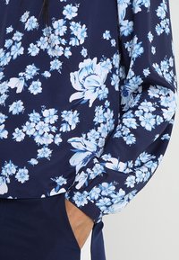 Blouse à manches longues en tissu bleu marine avec un motif floral en bleu clair et blanc. Texture douce avec une coupe décontractée aux poignets.