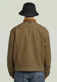 Veste en coton marron avec col en velours côtelé, couture visible et poches latérales, associée à un chapeau bucket noir, vue de l'arrière.