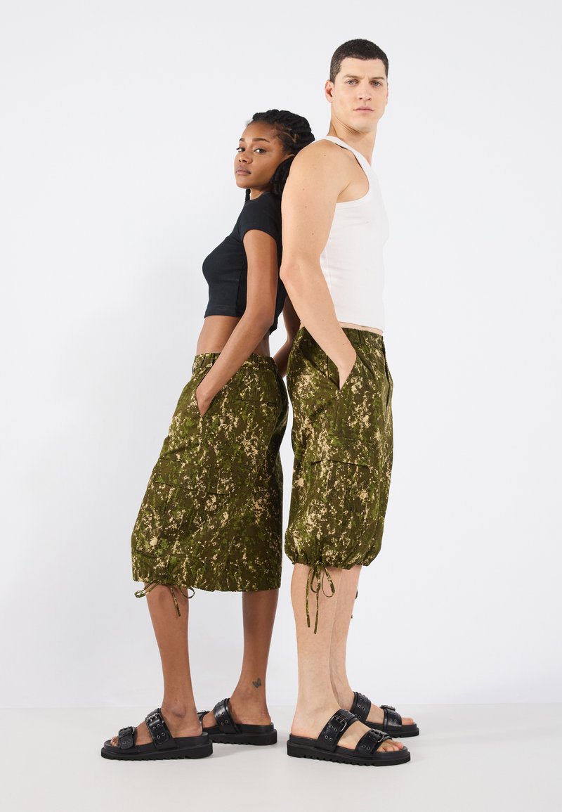 Gröna kamouflage shorts med sidofickor och dragsko i nederkant, tillsammans med en svart crop top och en vit linne. Svarta sandaler med spännen.