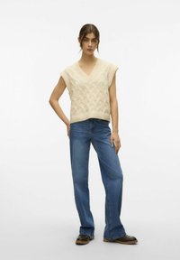 Ivore gebreide mouwloze top met een V-hals en een textuurpatroon, gecombineerd met blauwe flare jeans en bruine schoeisel, contra een effen achtergrond.