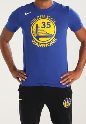Person iført en blå Golden State Warriors t-shirt med nummer 35 og sorte bukser, stående med hænderne på hofterne.