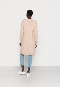 Beige stickad lång tröja med lös passform, rund halsringning och ribbade manschetter, ihop med ljusblå slitna jeans och vita sneakers.