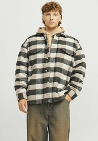 Camicia flanella a quadri in nero, bianco e grigio chiaro. Presenta due tasche sul petto, chiusure a bottone e una vestibilità comoda. Indossata sopra un felpa beige.