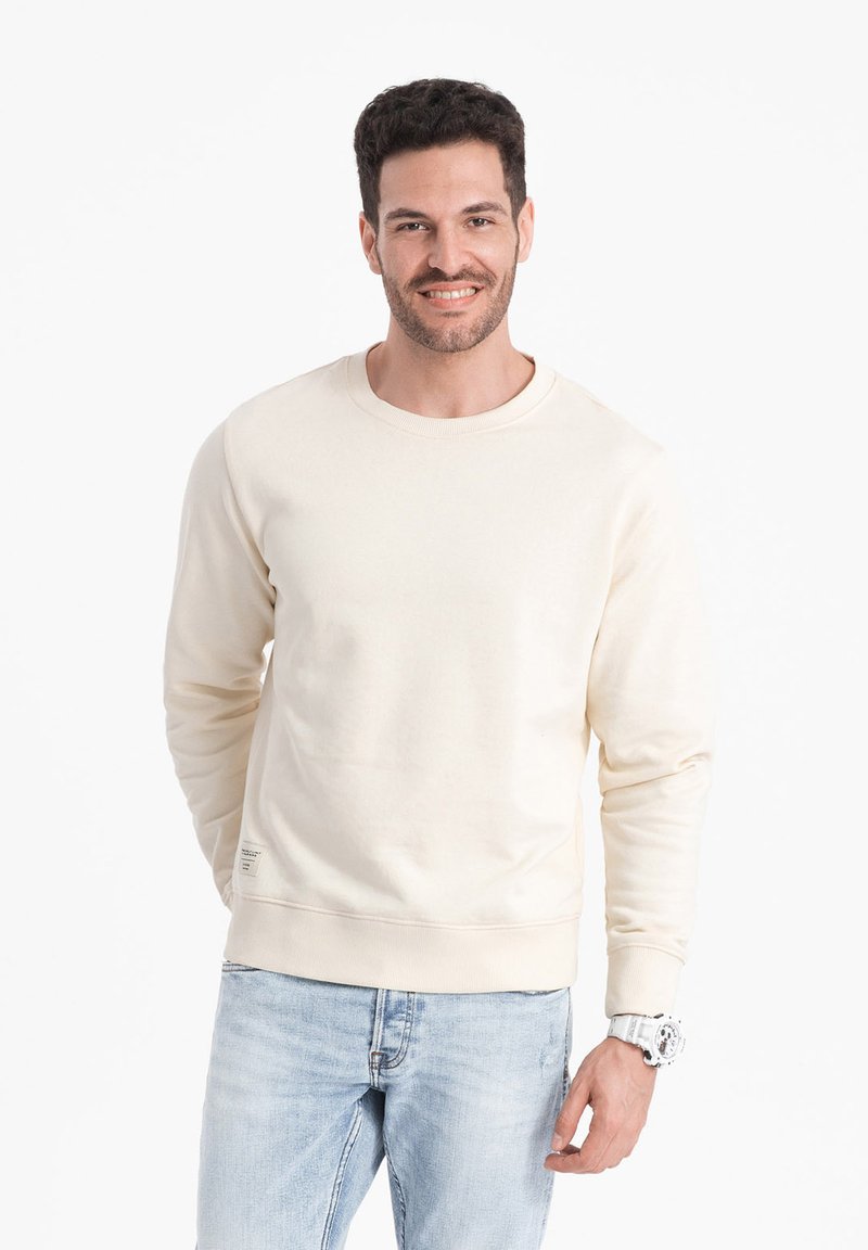 Sweatshirt di colore crema chiaro realizzato in tessuto morbido, con scollatura a girocollo, polsini e orlo a costine. Abbinato a jeans azzurri chiari e un orologio bianco.
