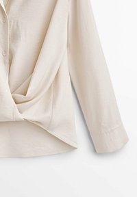 Blouse de couleur crème avec un design torsadé à la taille, manches longues et fermeture par boutons. Texture douce et tissu léger.