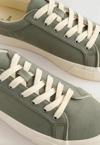 Baskets basses en toile vert olive avec des lacets crème et des semelles en caoutchouc, présentées côte à côte sur un fond clair.