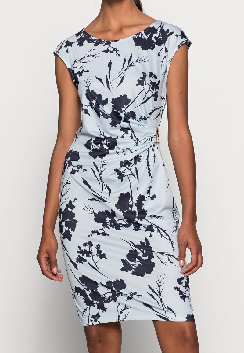 Femme portant une robe bleu clair mi-longue avec imprimé floral noir et détail froncé sur le côté droit.
