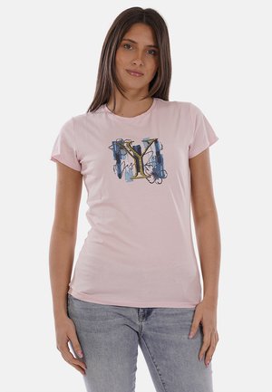 Donna con capelli lisci e scuri che indossa una maglietta rosa chiaro con un motivo decorativo a forma di "Y" e jeans azzurri, in piedi davanti a uno sfondo semplice.