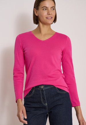Mujer que lleva una camiseta de manga larga de color rosa brillante con cuello en V y vaqueros azul oscuro, mirando ligeramente hacia su derecha y sonriendo suavemente.