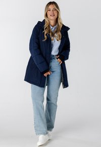 Marineblauwe gewatteerde jas met een capuchon, rits aan de voorkant en zijzakken, over een lichtblauw gestreept shirt, gecombineerd met lichtblauwe broek en witte sneakers.