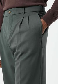 Donkergroene geplooide broek van een gladde stof, met een voorzak en een bruine knoop in de taille.