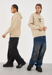 To personer iført beige hættetrøjer og løstsiddende jeans står ryg mod ryg på en neutral baggrund, den ene vender sidelæns, den anden ser ned.