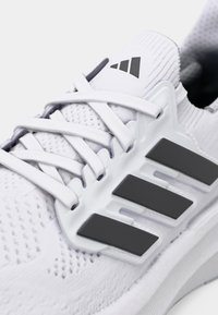 Valkea urheilukenkä, jossa on verkkokuosi ja harmaat korostukset. Sivulla näkyy selkeä kolmen raidan logo. Nauhallinen malli.