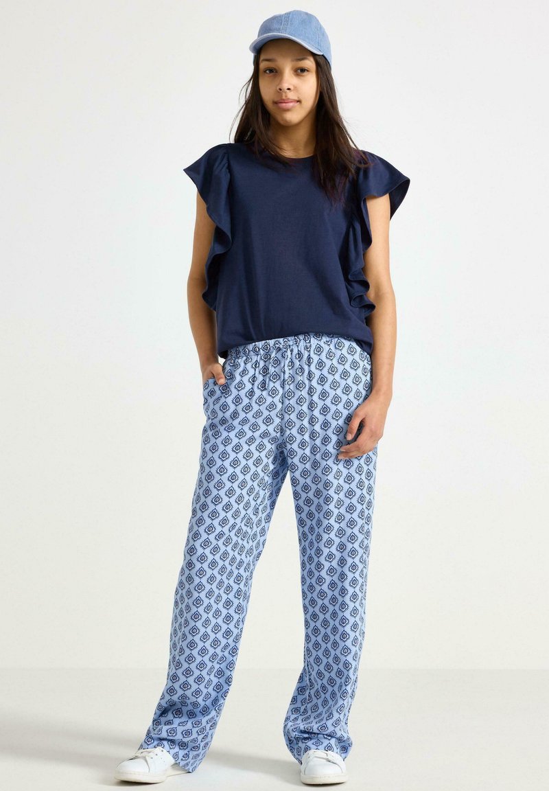 T-shirt blu navy con maniche a volant, abbinato a pantaloni a fantasia blu chiaro con forme a diamante, completato con un cappellino blu chiaro e sneakers bianche.