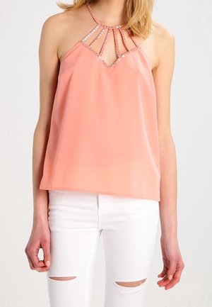 Vrouw draagt een mouwloze perzikkleurige haltertop met paillettenbandjes en witte gescheurde skinny jeans tegen een effen achtergrond.