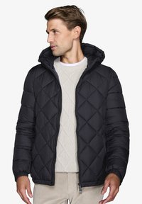 Sort polstret jakke med en quiltet mønster, der har en lynlås og hætte. Båret over en lys cremefarvet sweater med et tekstureret design.