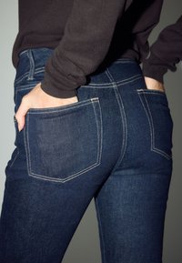 Sötétkék farmernadrág, testhezálló fazonú, hátsó zsebekkel és kontrasztos világosbarna varrással, neutrális háttér előtt elhelyezve.