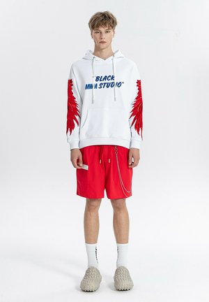 Joven con sudadera blanca con capucha y diseños de alas rojas en las mangas y texto azul, pantalones cortos rojos con cadena, calcetines blancos y zapatos beige.