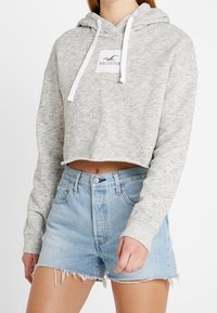 Krátká mikina s kapucí v světle šedé barvě, vyrobená z měkké látky s bílou nášivkou loga. Kombinována s džínovými šortkami s vysokým pasem a třepenými lemy.
