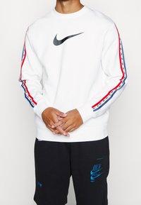 Vit träningströja med svart Nike-logotyp, röda och blå randiga detaljer på ärmarna, ribbstickade muddar och rund halsringning. Bärs med svarta shorts.