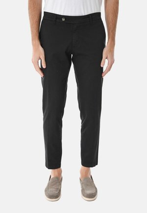 Uomo che indossa pantaloni neri corti, camicia bianca e scarpe slip-on grigie, in piedi contro uno sfondo semplice.