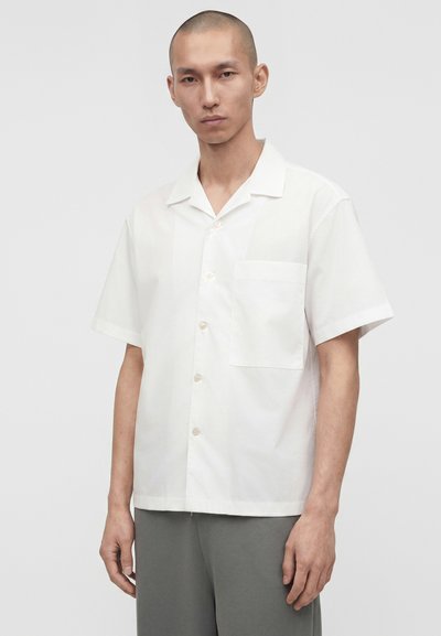 Νέος άνδρας με κοντά μαλλιά, που φοράει απλό λευκό κοντομάνικο πουκάμισο και γκρι παντελόνι, σε ανοιχτό γκρι φόντο.