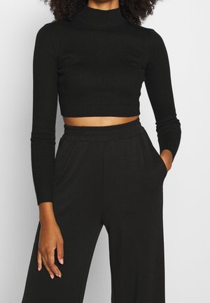Modèle portant un haut court noir à col roulé côtelé et un pantalon noir taille haute, une main dans une poche.