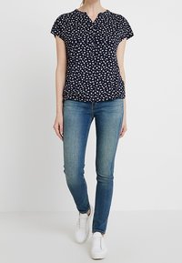 Kvinna klädd i en marinblå mönstrad kortärmad blus, åtsittande blå jeans och vita sneakers, stående mot en enkel vit bakgrund.