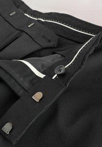Pantaloni negri cu închidere prin fermoar și nasture, detalii de cusătură albă și accente din metal. Textura fină a țesăturii este vizibilă.