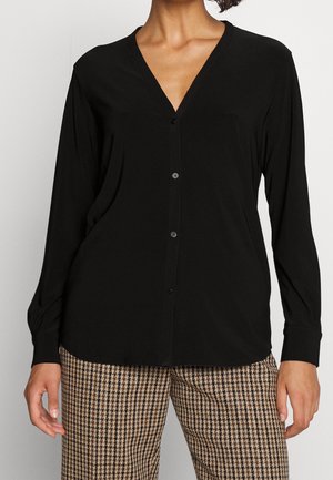 Kobieta w czarnej, długorękawowej bluzce na guziki i brązowych spodniach w wzór pepitki, stoi z rękami swobodnie opuszczonymi wzdłuż ciała.