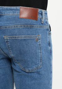 Sinised teksad, millel on nahast tagumine silt "BOSS", klassikalise taskukujunduse ja nähtavate õmbluste detailidega.