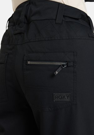 Pantalons noirs en tissu lisse, dotés d'un design matelassé, d'une poche zippée et d'un patch logo Roxy au dos. Des coutures définies sont visibles.