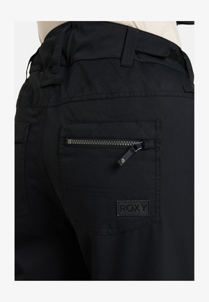 Schwarze Hose aus glattem Stoff mit einem gesteppten Design, Reißverschlusstasche und einem Roxy-Logo-Patch auf der Rückseite. Sichtbare Nähte sind vorhanden.