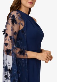 Navyblauwe jurk met een één-schouder ontwerp en een doorschijnende overlay met geborduurde bloemen en bladeren op de mouw. Soepele stofstructuur.