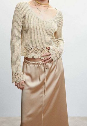 Crop top en maille beige clair avec bordure en dentelle, associé à une jupe en satin beige brillante taille haute, nouée avec un fin nœud de corde.