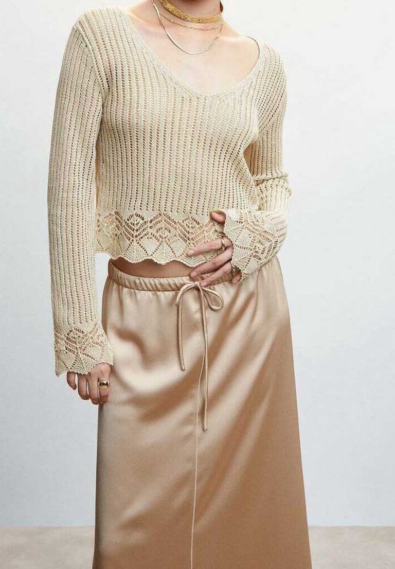 Crop top en maille beige clair avec bordure en dentelle, associé à une jupe en satin beige brillante taille haute, nouée avec un fin nœud de corde.