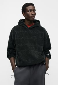 Σκούρο πράσινο φούτερ με κουκούλα από υφασμάτινη υφή, με τσέπη καγκουρό και διακριτικό μοτίβο με λογότυπο σε μορφή γραφής. Συνδυάζεται με γκρι παντελόνι.