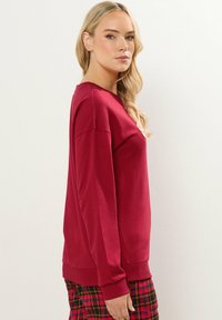 Sudadera roja de manga larga con escote redondo y puños acanalados. Hecha de un tejido suave, combinada con pantalones de patrón de cuadros.