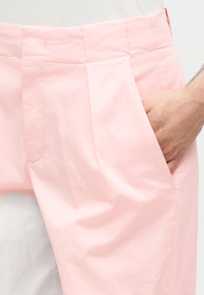 Main dans la poche d’un short rose clair sur mesure porté avec un haut blanc, montrant les détails de la taille et des plis.