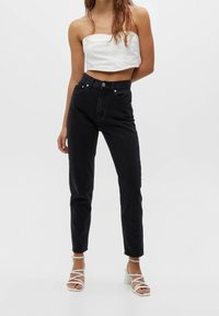 Femme portant un jean noir taille haute, un crop top blanc sans bretelles et des sandales à talons blocs à lanières blanches, debout sur un fond clair uni.