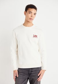 Felpa crema con maniche lunghe e collo rotondo, con un logo rosso "Lee" sul petto. Abbinata a jeans grigio scuro.