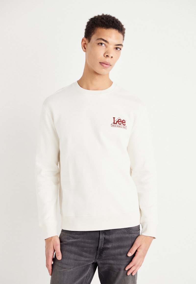 Felpa crema con maniche lunghe e collo rotondo, con un logo rosso "Lee" sul petto. Abbinata a jeans grigio scuro.