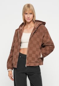 Giovane donna che indossa un jacket a quadri in velluto a coste marrone con cappuccio, un crop top beige e pantaloni scuri, in piedi contro uno sfondo bianco.