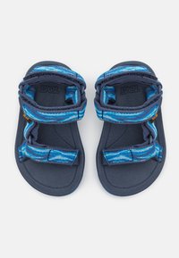 Blå og marineblå sandaler med to stropper med et strukturert, bølgete mønster og justerbare fester. Strukturert fotseng og gummisåle.