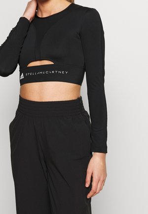 Kobieta ubrana w czarny krótki top z długim rękawem i wycięciem oraz napisem "Adidas Stella McCartney", w połączeniu z czarnymi spodniami z wysokim stanem.