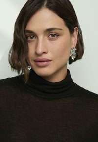 Model w wearing a black, dopasowany golf. Wyraźne srebrne kolczyki w kształcie liścia. Gładka skóra i średniej długości falowane brązowe włosy.